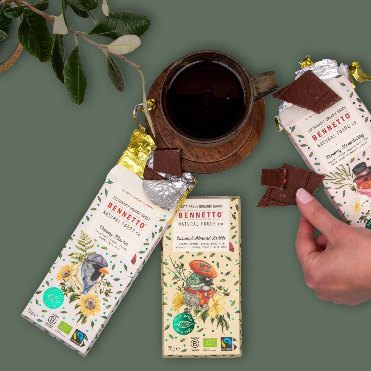 Three Bennetto chocolate bars with illustrated wrappers are arranged on a muted green background, alongside a dark cup of coffee, a small plant, and a hand holding a broken piece of chocolate.