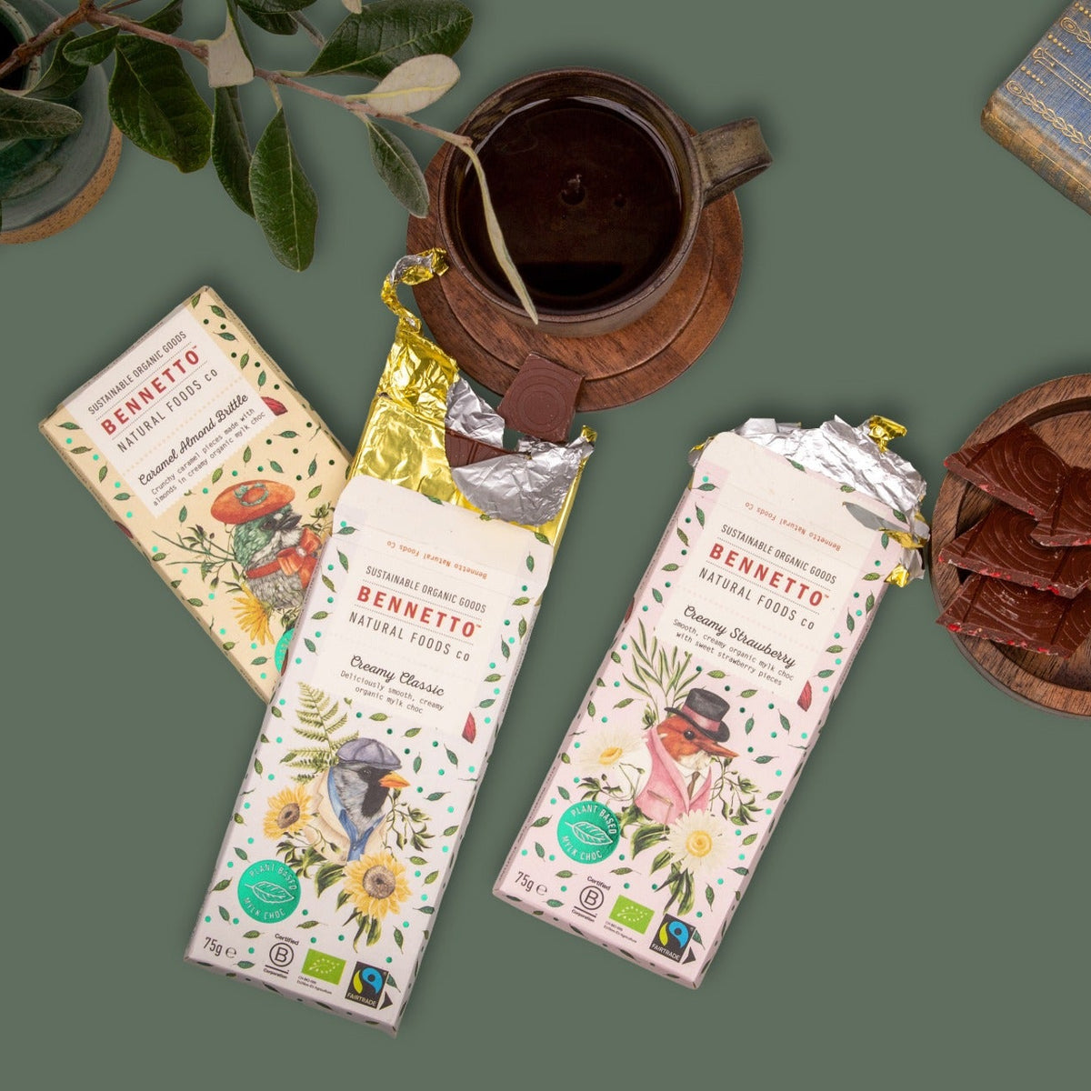 Two unwrapped Bennetto chocolate bars and one wrapped bar from the brand, arranged in a styled flat-lay with a ceramic mug, a book, and a wooden dish on a muted green background.