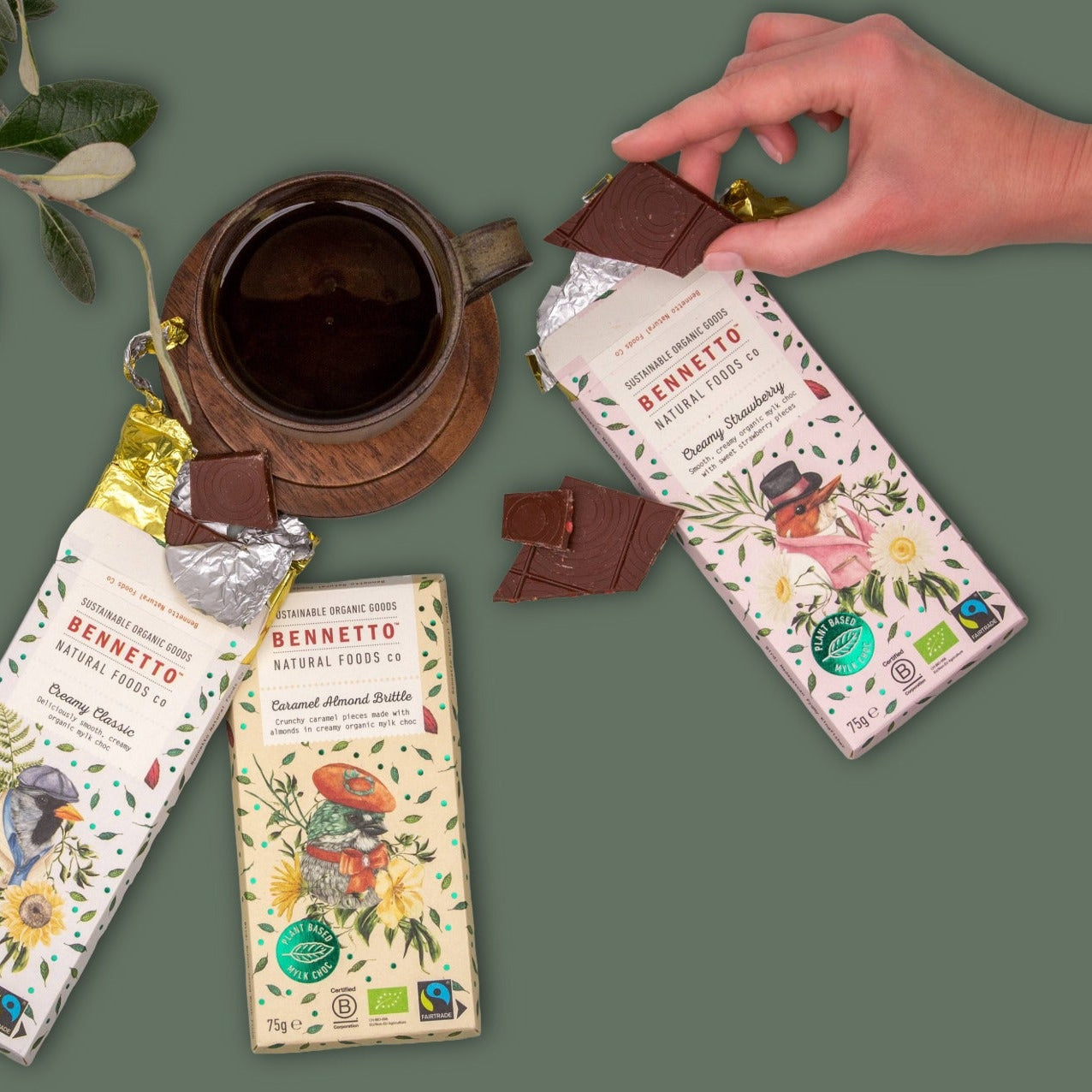 Three bars of Bennetto chocolate with illustrated pastel-colored wrappers are arranged on a muted green background alongside a dark ceramic cup and a hand holding a piece of chocolate. The wrappers feature botanical illustrations and text including 'BENNETTO' and 'Natural Food Co'.