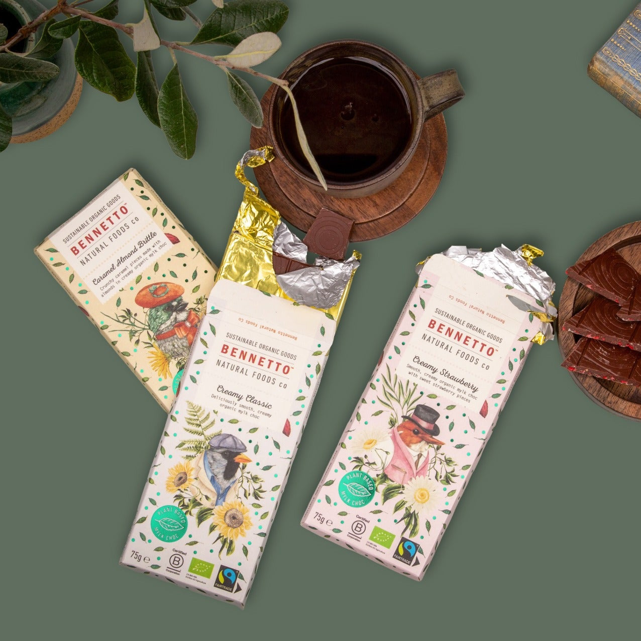 Two unwrapped Bennetto chocolate bars and one wrapped bar from the brand, arranged in a styled flat-lay with a ceramic mug, a book, and a wooden dish on a muted green background.