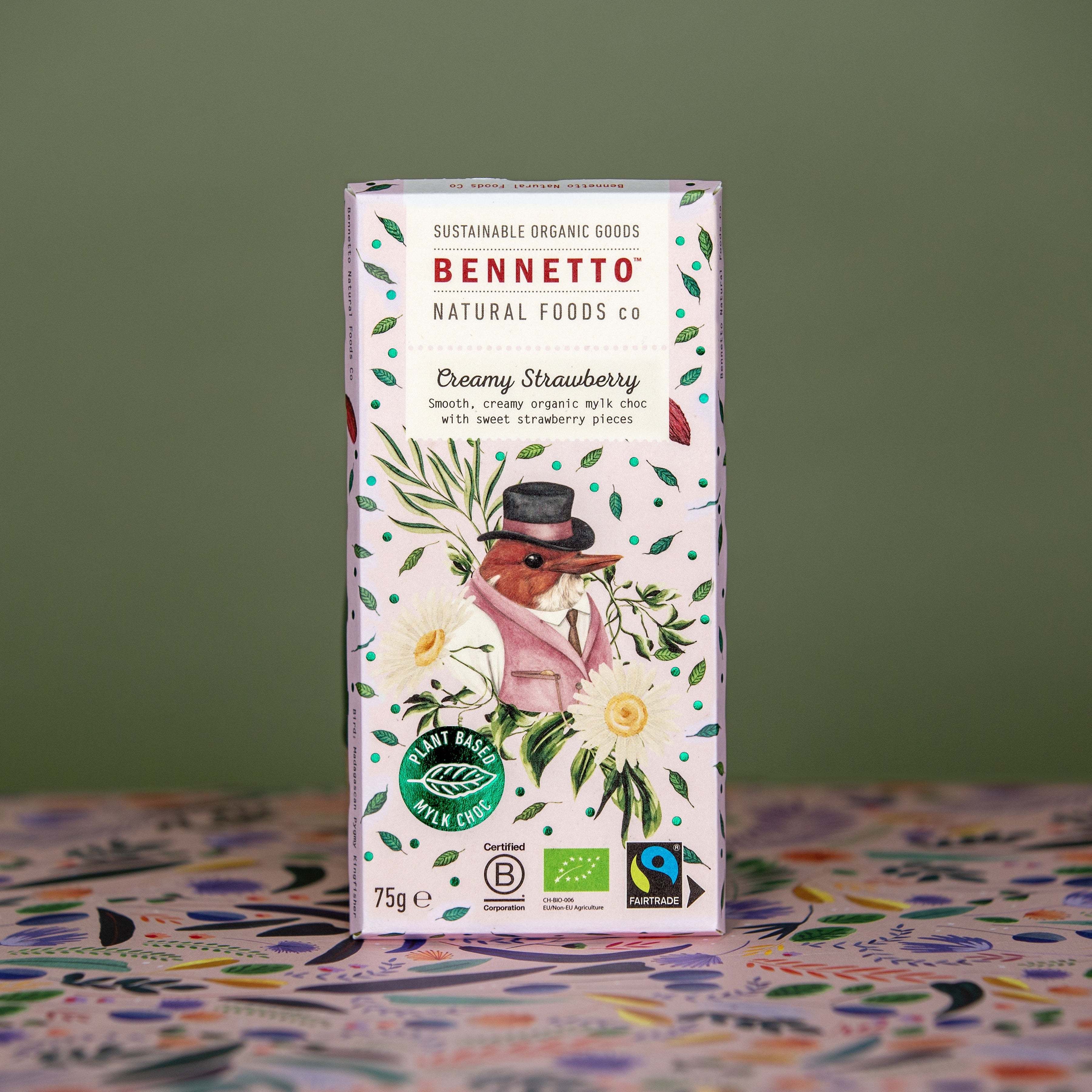 Bennetto Creamy Strawberry Mylk Chocolate standing upright on a green khaki background with patterned floral paper on the bottom. A 75g bar with bcorp, organic and fairtrade certifications. A light pink with a bird and florals on the front packaging.